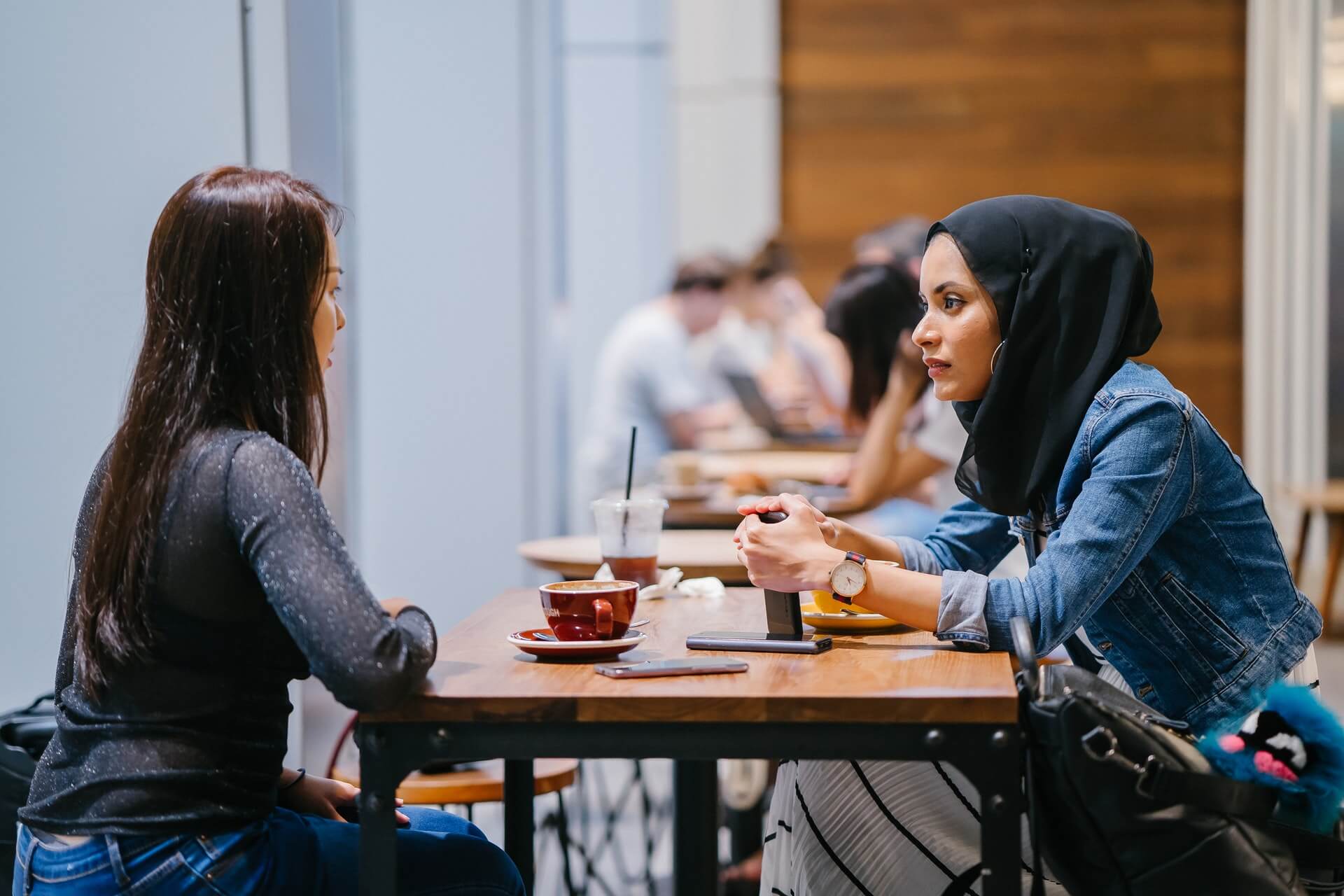 Two women sit on either side of a wooden table having coffee together. One woman is wearing a black headscarf while the other woman has long brown hair which is down