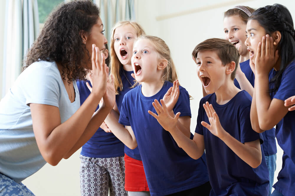 kids enjoying a drama class wearing blue shirts with their hands raised and their mouths open and their eyes wide with a look of surprise. Their teacher is wearing a light blue shirt and she is standing facing them with her hands up as well.