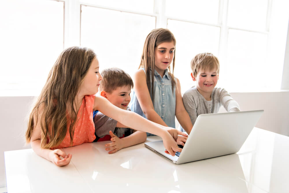 young children gathered around a silver laptop sitting on a white table they are all pointing and touching it there is a large wall of windows that is all white behind them