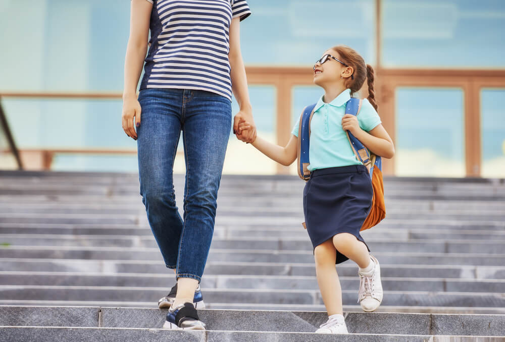 parent wearing a blue and white striped shirt and blue jeans and sneakers is holding the hands of her child child while they are walking down the stairs outside