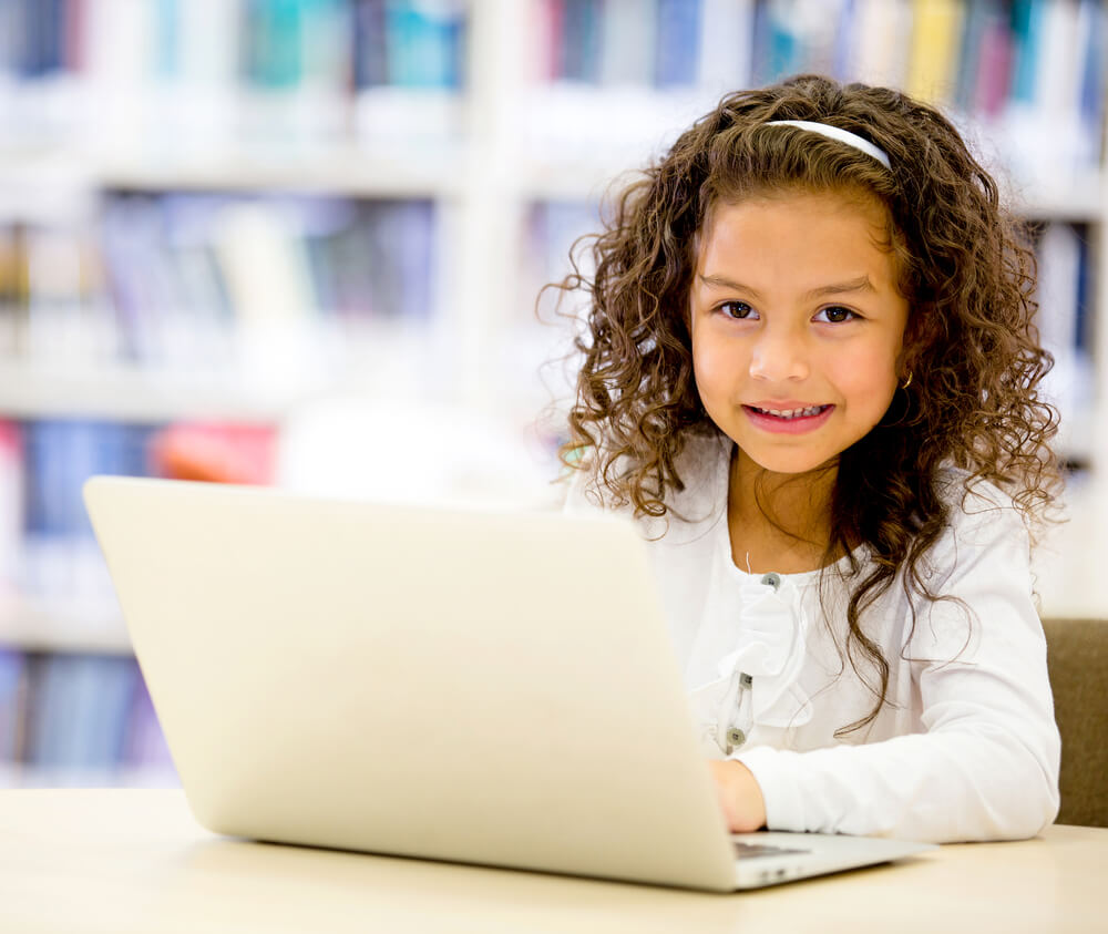 A little girl with brown curly hair and a white headband and a white shirt is sitting in front of a bookshelf and using a silver laptop
