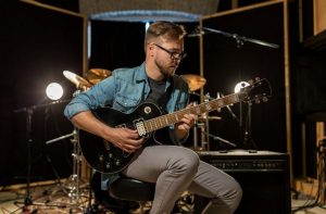 A man wearing grey pants and a blue shirt holds a black electric guitar in a recording studio in front of a drum set
