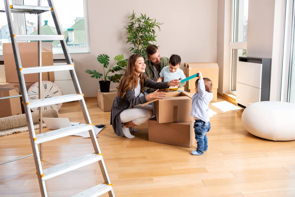 A mother and a father and two young sons are sitting in the middle of the room around packing boxes after moving into a new home. They are in the middle of unpacking the boxes together. There are some plants behind them as well as more boxes. In the foreground of the photo is a tall ladder.
