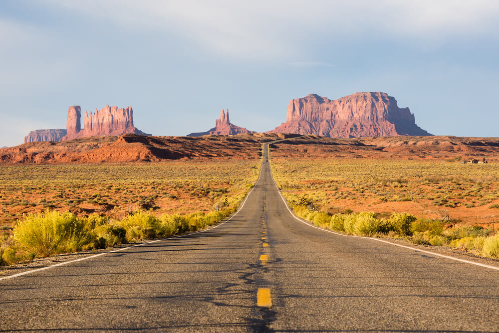 A long black road that disappears into the horizon at monument valley with mountains and rock formations far out in the distance