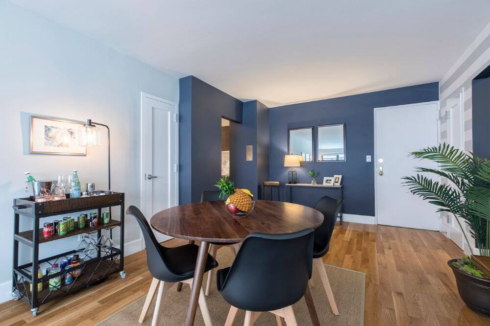 A furnished and equipped dining area in an NYC apartment managed by Blueground. There is a round wooden table with black chairs around it and a black bar cart off to the side. There is a potted plant to the right and a dark blue wall with framed photos in the background