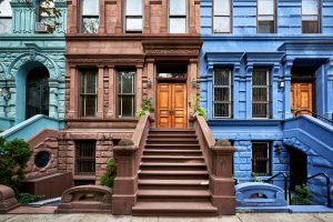 multicolored brownstones show the various building facades of New York City. In the center is a brown building with stairs and a wooden door with two blue buildings on either side.