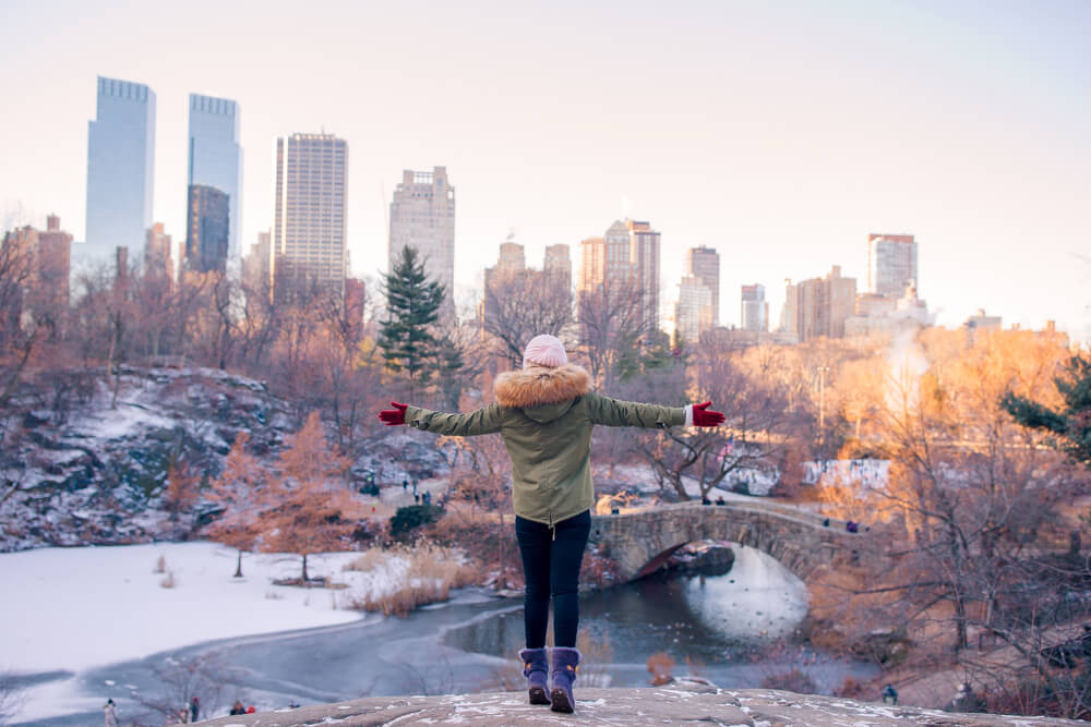 During Winter in NYC Central Park a girl wearing a green jacket and red gloves and black leggings and purple boots is standing with her arms outstretched looking towards the buildings and bridge in front of her