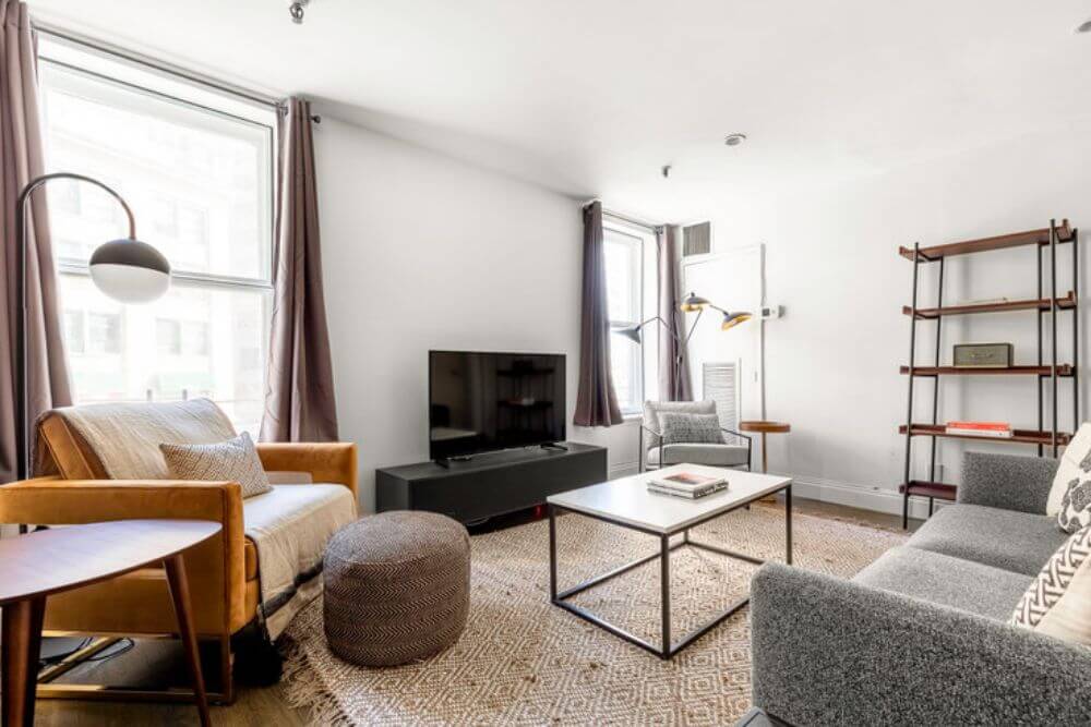 A furnished apartment in NYC managed by Bluegrond. There are many browns and neutral tones. The couch is grey and sits across from a TV on a TV stand a white coffee table. There is a brown leather chair in the corner with a white throw blanket on top.
