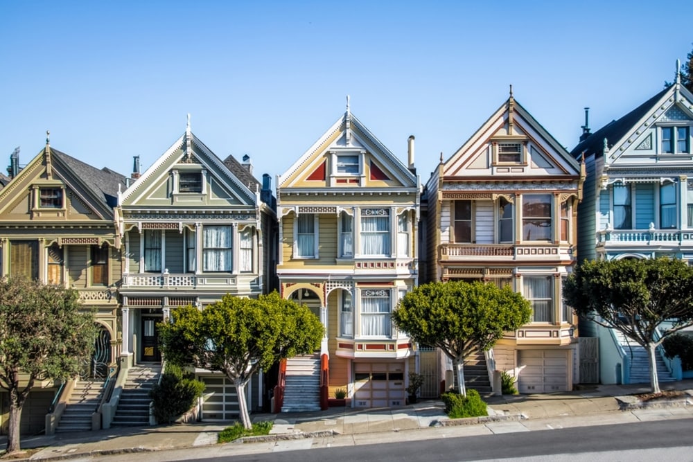 The multicolored houses in a row in San Francisco that is called the Painted Ladies. The street goes up at an angle and there are trees in front of the homes with a blue sky behind them