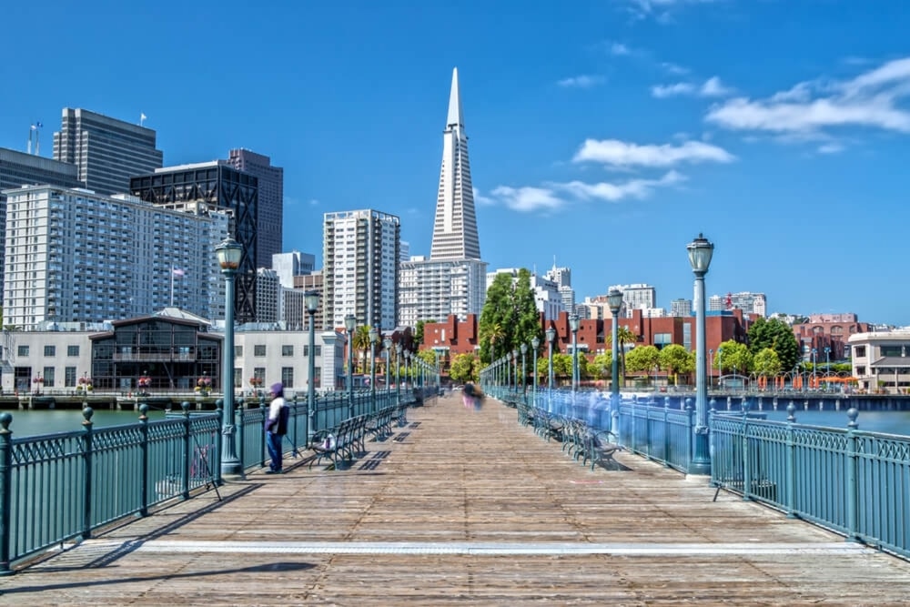 A wooden pier in San Francisco with water on either side and the Transamerica Pyramid building at the end. There is a bright blue sky with a small white cloud on the right side.