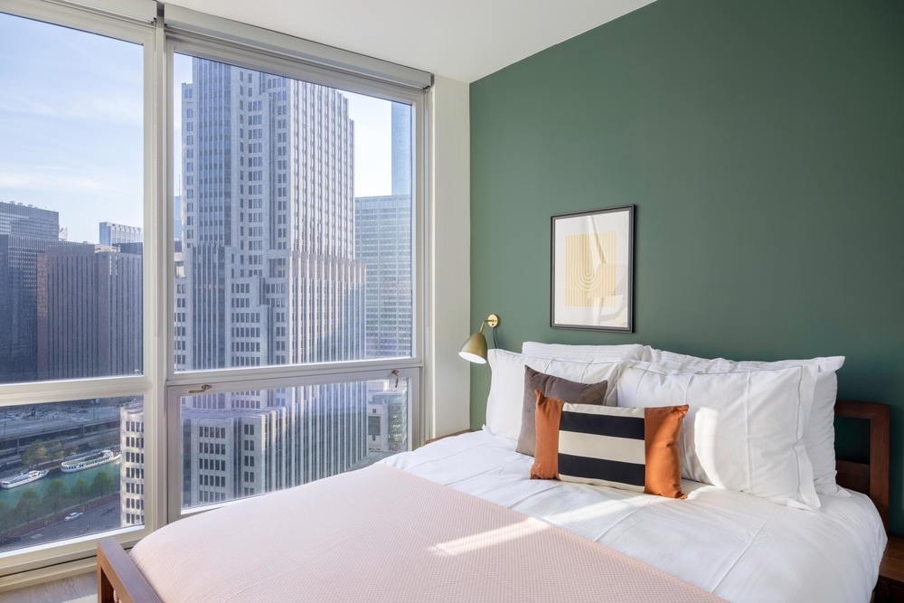 A large wooden bed frame with white sheets, a pink blanket and multicolored pillows sits in front of a dark green accent wall. The bed sits next to a large glass window with a view of some tall buildings