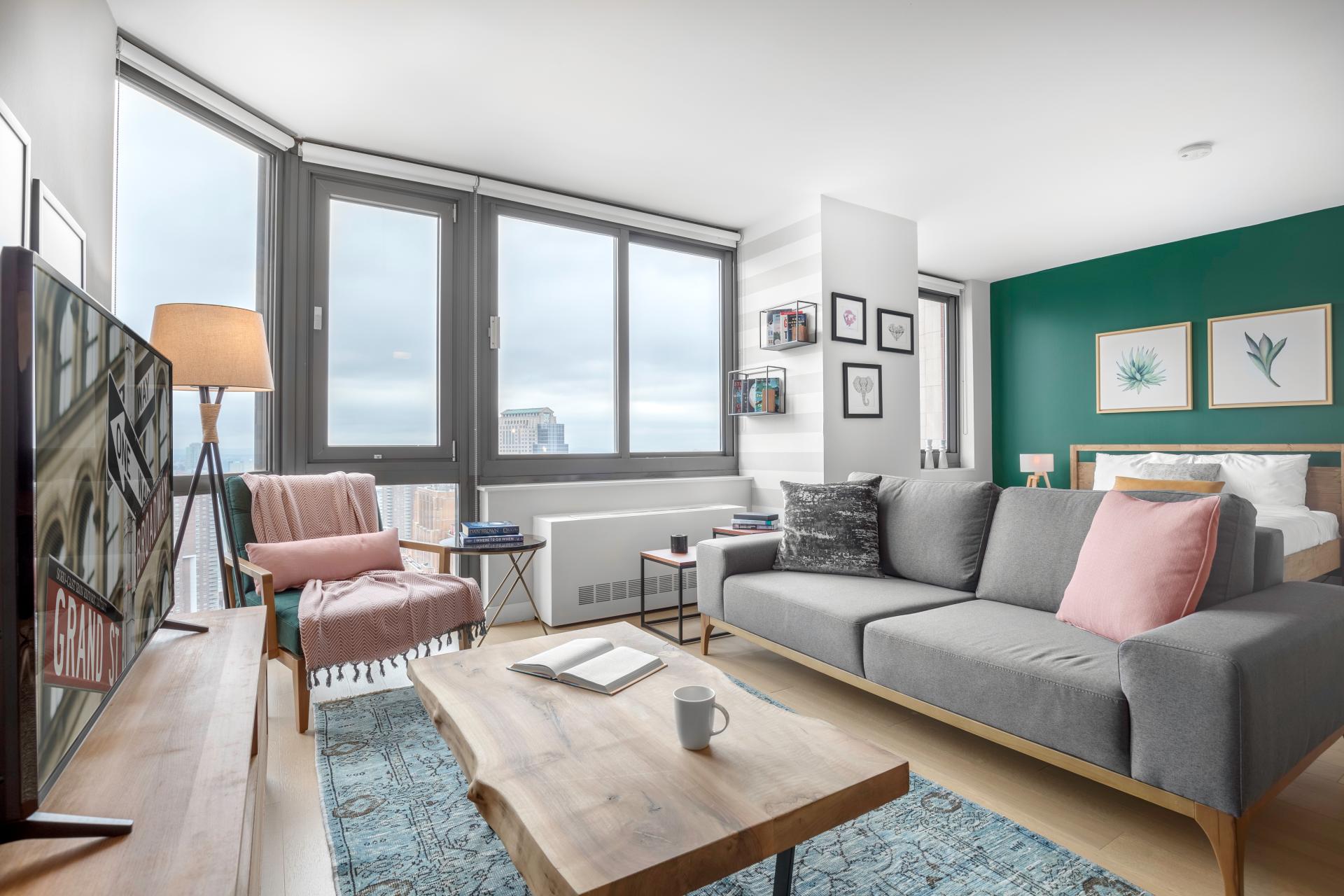 A studio apartment that has been furnished and managed by Blueground. There is a grey couch in the center of the room with a wooden coffee table in front, facing the TV. Behind the couch is a large bed and in the corner is an armchair