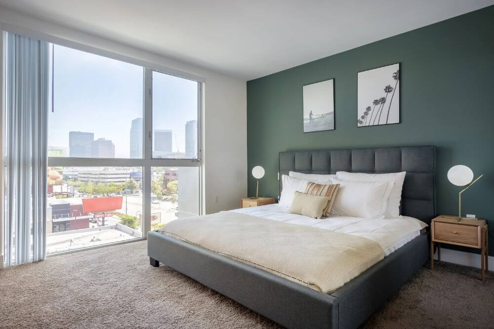 A furnished bedroom in Westwood Los Angeles. There is a dark green accent wall and a large black double bed next to a large glass window with a view of tall buildings and trees. There are two framed black and white photos above the headboard and a nightstand with a lamp next to the bed.