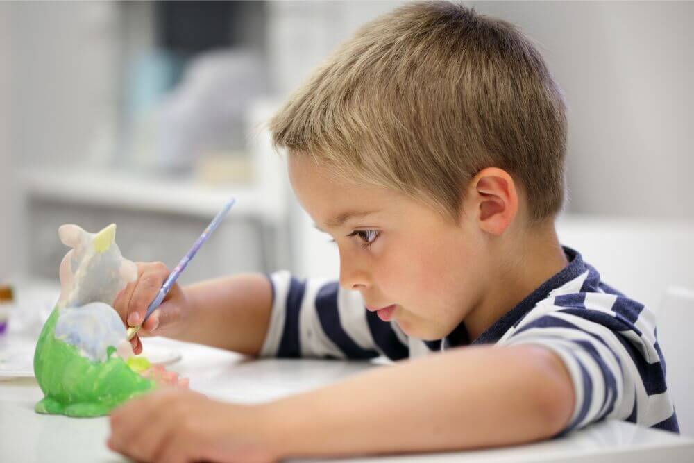 A boy with blonde hair wearing a white and blue striped shirt is holding a paintbrush in his hand and painting the color green onto a piece of ceramic.