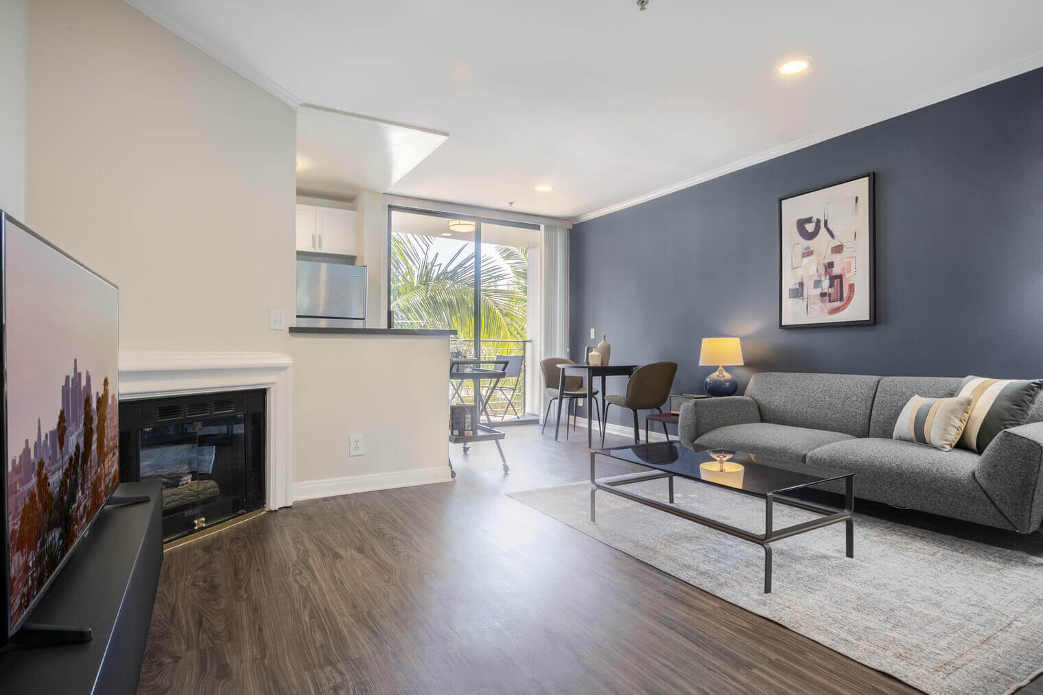 A furnished apartment in Brentwood Los Angeles. There is a fireplace next to a large TV on a large black TV stand across from a grey couch and a marble coffee table. In the back of the room there is a large window with a palm tree outside.