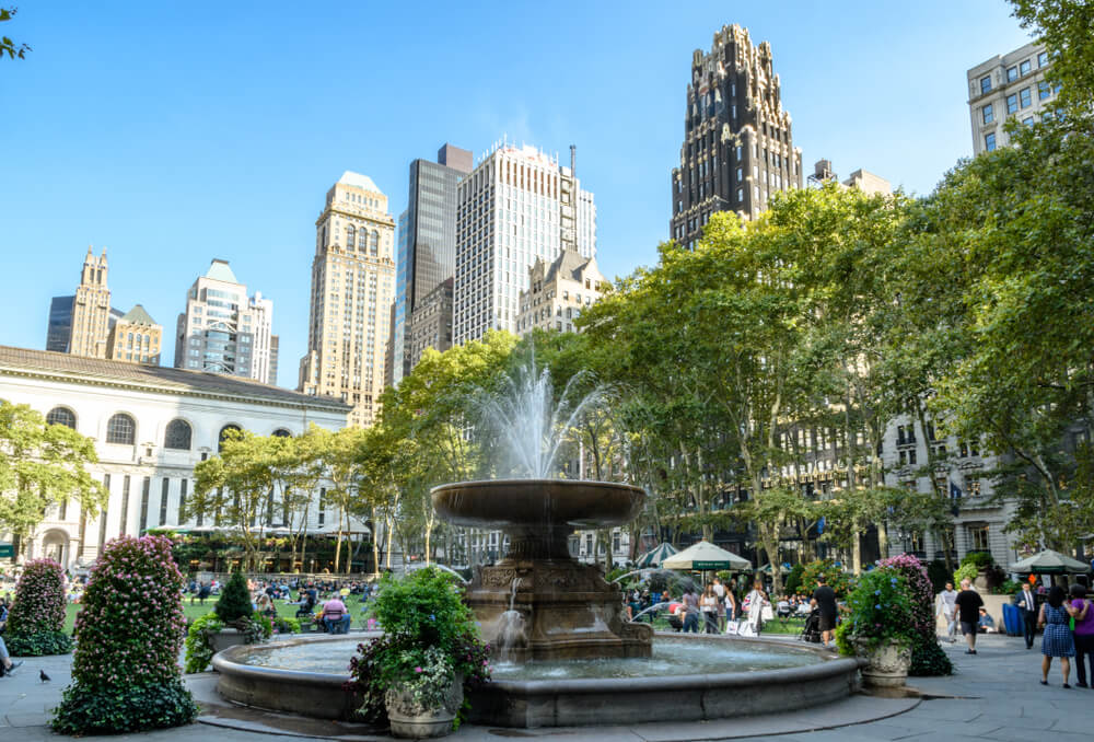 A large fountain in Bryant Park with greenery around and tall high rise buildings in the background under a clear blue sky
