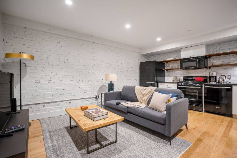 A furnished apartment in Boston with a brick wall painted white and a grey couch and a wooden coffee table. There is a fully-equipped kitchen area behind the couch with all the necessary appliances.