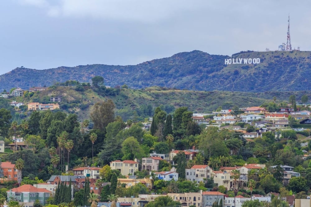 Panoramic view of Hollywood Hill with the hollywood sign and the antenna behind it overlooking smaller hills with lots of large houses and trees 
