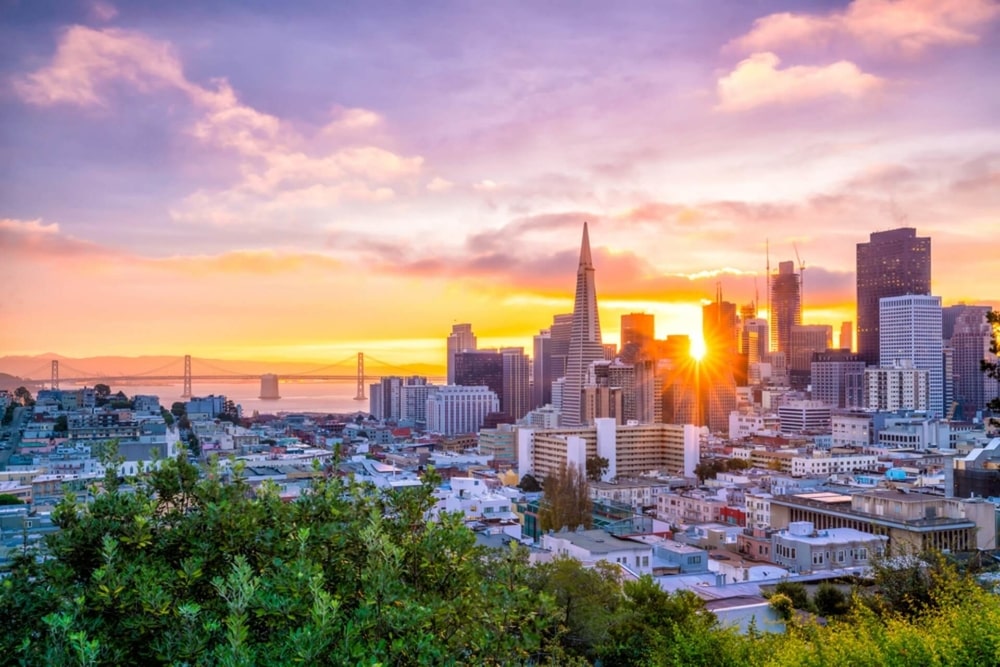 sunrise over the skyline of San Francisco with the rays of sun coming through the tall buildings. There is faint image of the golden gate bridge in the background on the left side