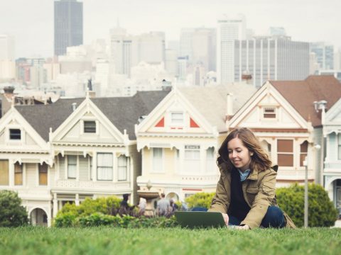 housing San Francisco Young girl sitting on a lap top on a lawn in front of traditional San Francisco style housing