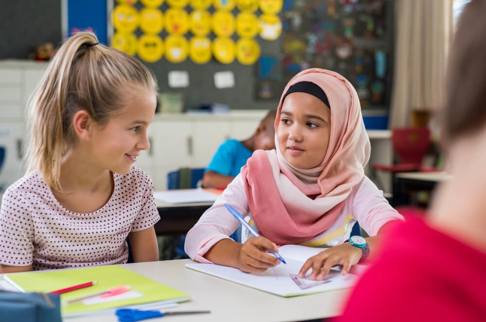 A blonde girl is at school sitting next to another young girl wearing a pink headscarf. The girl with the headscarf is holding a blue pen in one hand and a ruler in the other on an open white notebook.