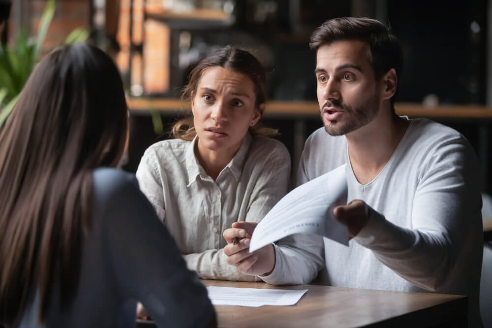 A man and a woman are sitting across a wooden table from a woman with long brown hair. They are arguing. The man is holding up a paper with one hand and a pen in the other hand.