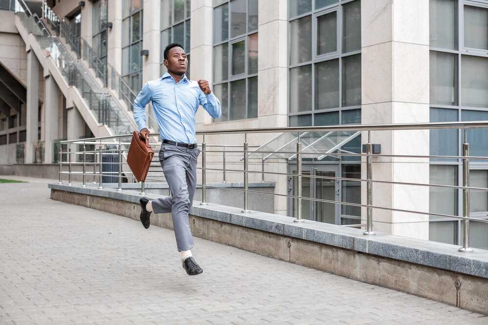 A man wearing a blue shirt and grey pants is running through the city while he is holding a brown leather briefcase