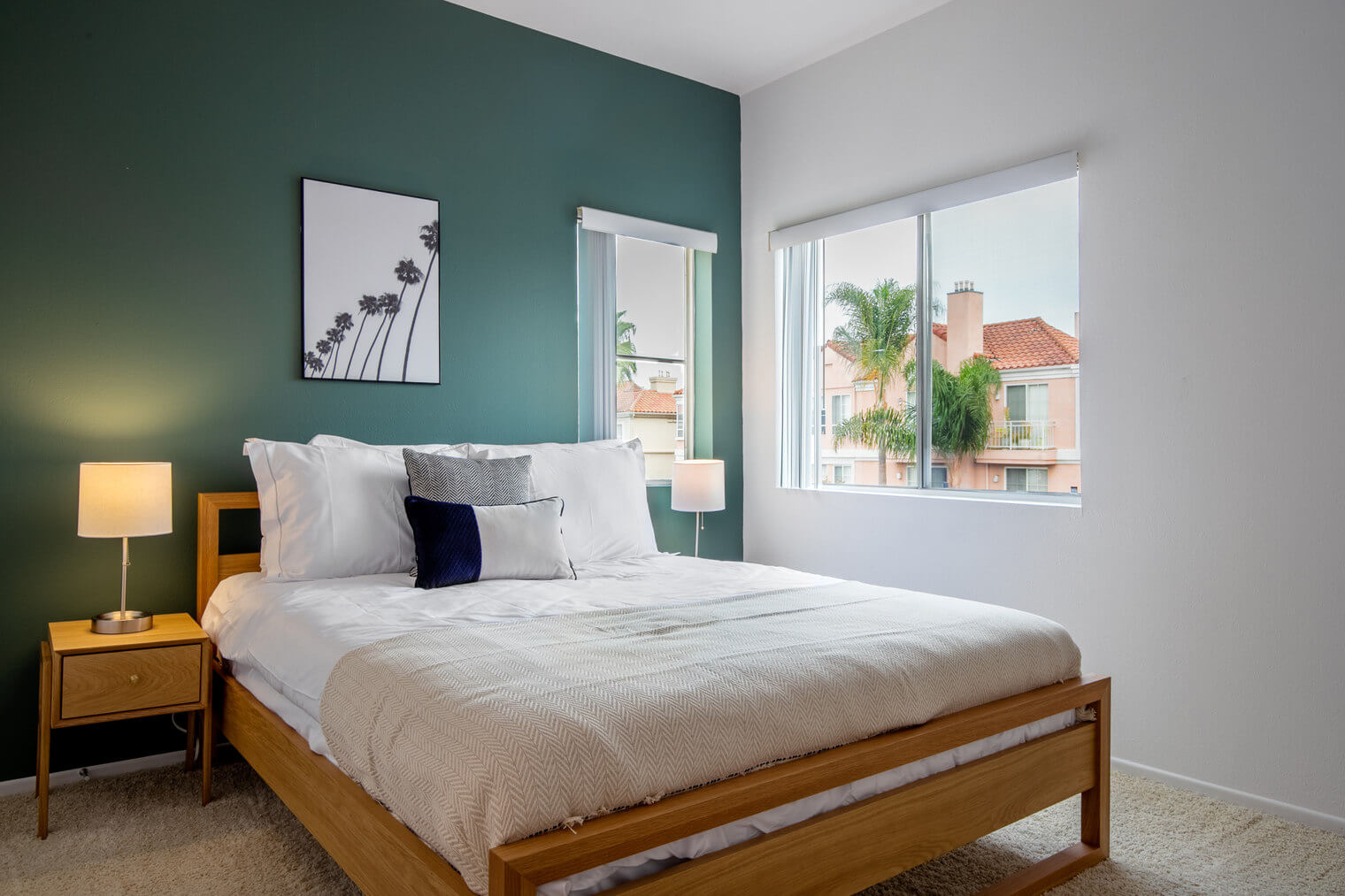 A dark green accent wall with a large wooden bed with lots of pillows and a light yellow blanket. Above the pillows is a framed black and white photo of plam trees. On the side of the bed is a window with a view of a pink house and large palm trees.