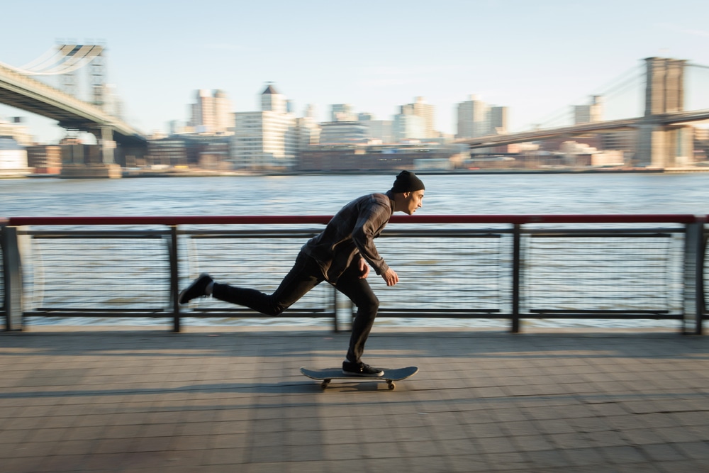 A man wearing black clothes and a black hat is skateboarding in new york city there is water on his left side and across from that are many buildings in the city skyline