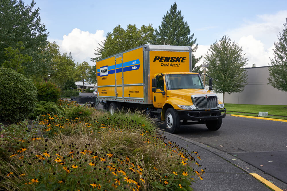 A large yellow penske truck driving down the highway next to a grassy area with flowers and trees