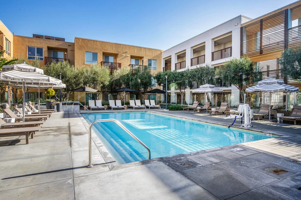 A large outdoor pool in the common area of an apartment building in Play Vista California. There are many pool chairs and umbrellas with trees behind them. The buildings are white and bright yellow.