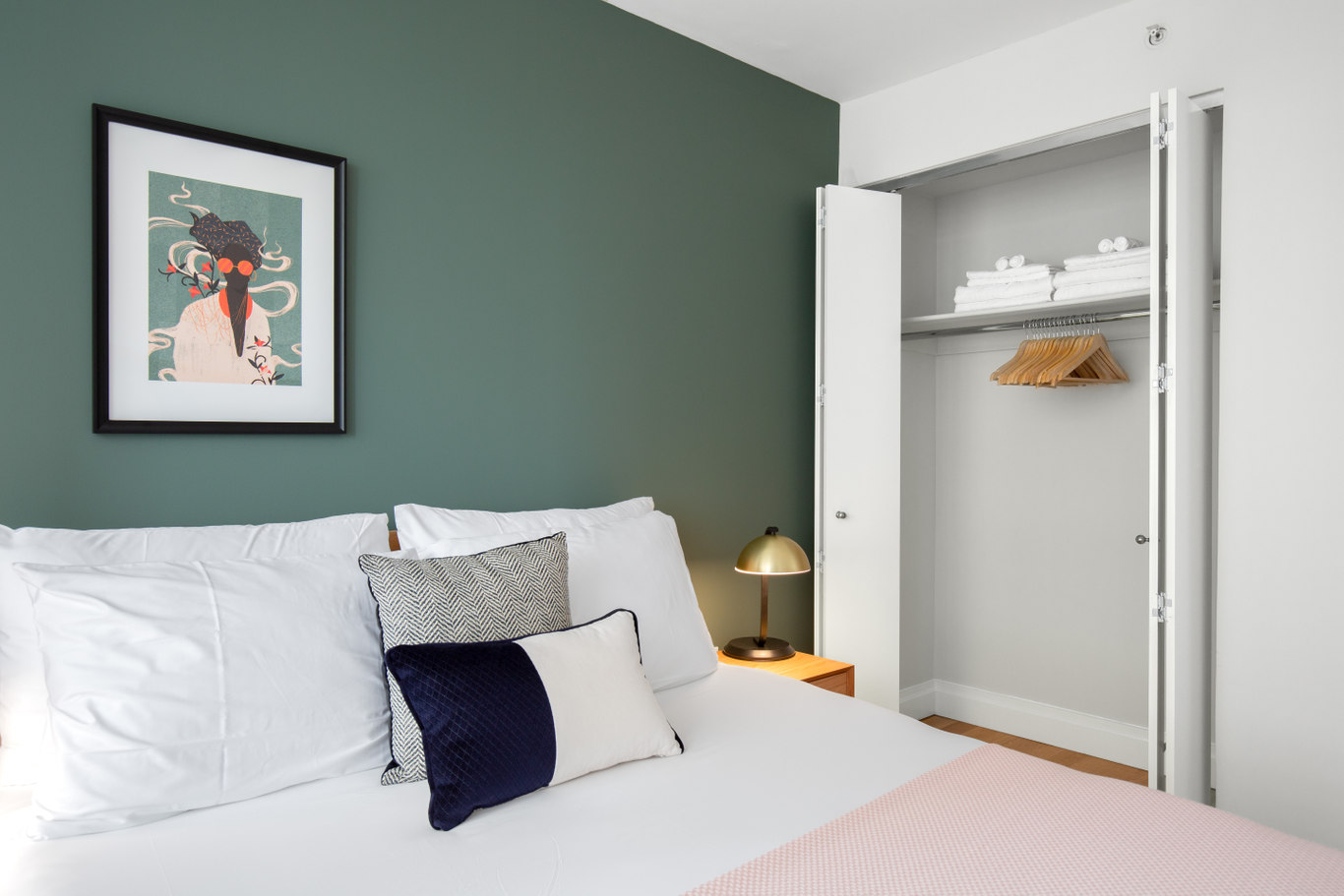 A small bedroom with a green wall with a painting in a black frame above a large bed with white linens. On the right side there is an open closet with wooden hangers and white towels
