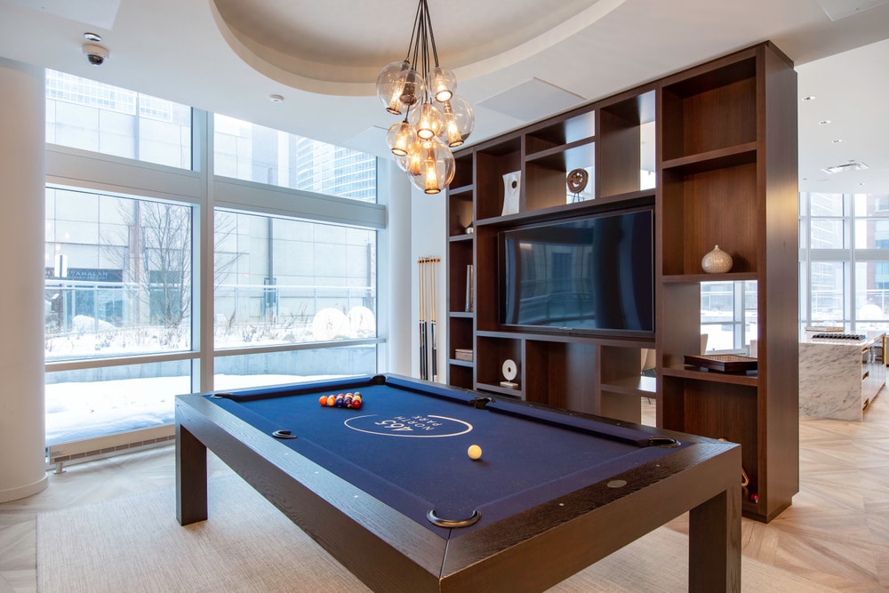 A large wooden pool table next to a large wooden bookshelf with a T.V. in the center. There are many decorative items around the shelves and on the right side is a large glass window