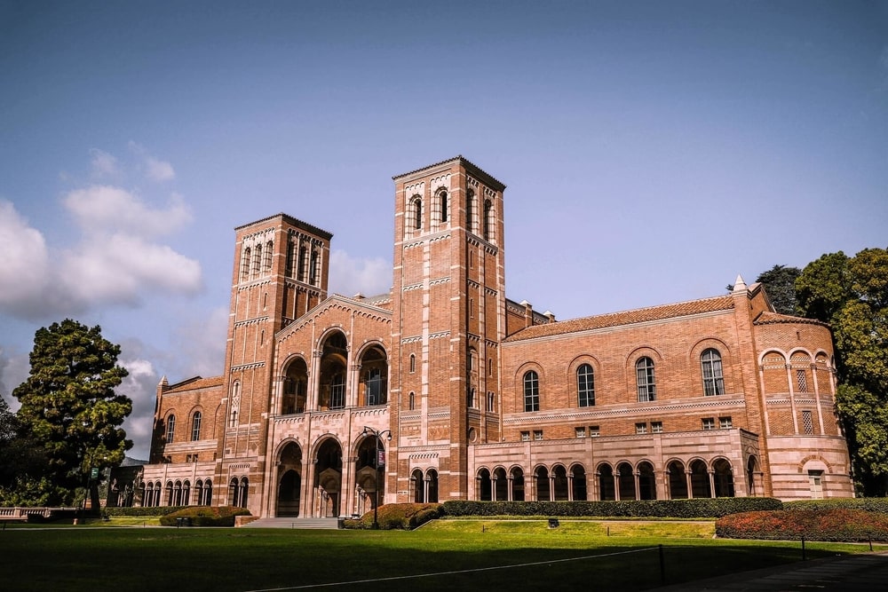 A facade of a large red brick building in UCLA campus with a green lawn in front and a blue sky with some white clouds above on the left side