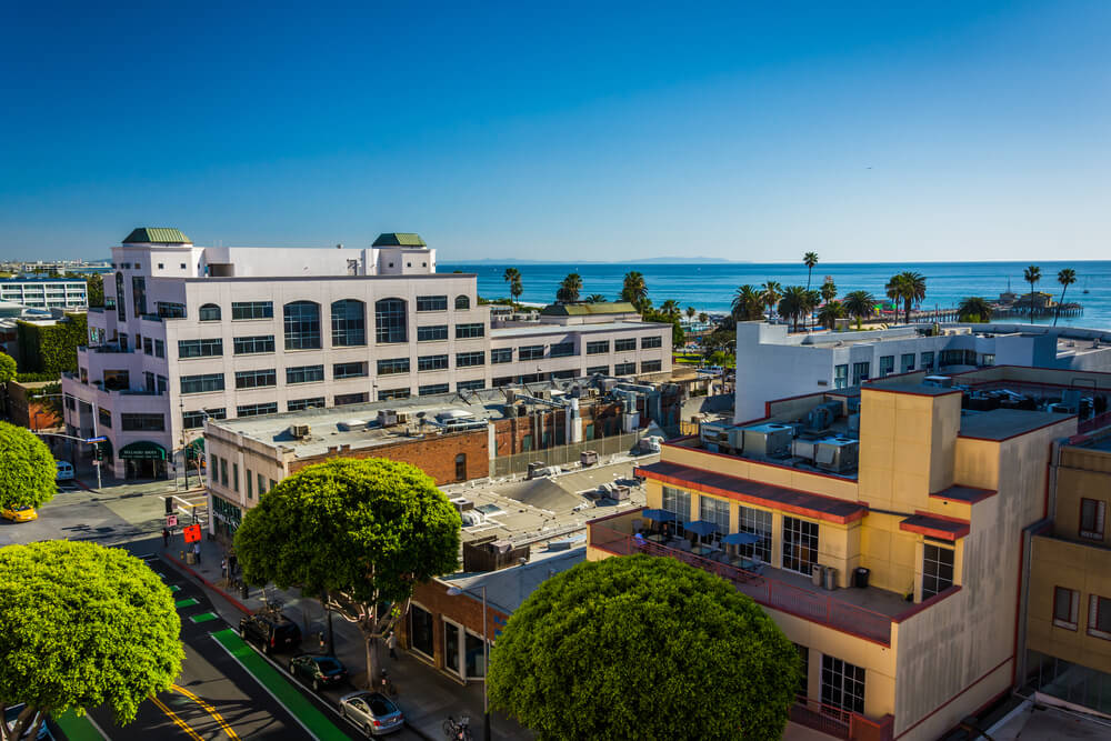 An overview of large buildings and trees in Santa Monica in Los Angeles California. There is a view of the Pacific Ocean in the background underneath a clear blue sky.
