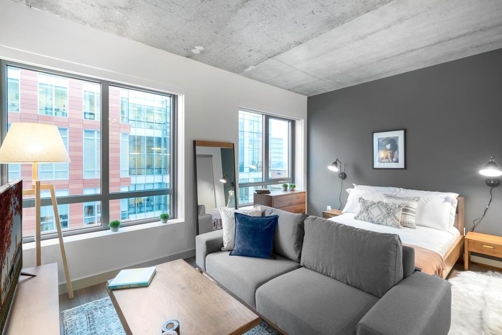 A studio apartment in Boston with a grey sofa and a wooden coffee table in front. Behind the grey couch is a wooden bed and a dark grey accent wall.