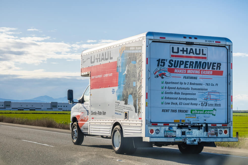 A large white uhaul moving truck is driving down the highway with a blue sky above
