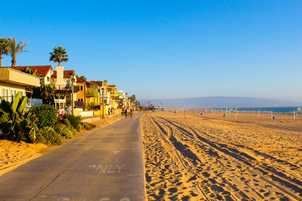 A long row of large upscale houses on the left side and a cement sidewalk in front of them with a large stretch of sandy beach on the right side. THere is a mountain in the distance in the back.