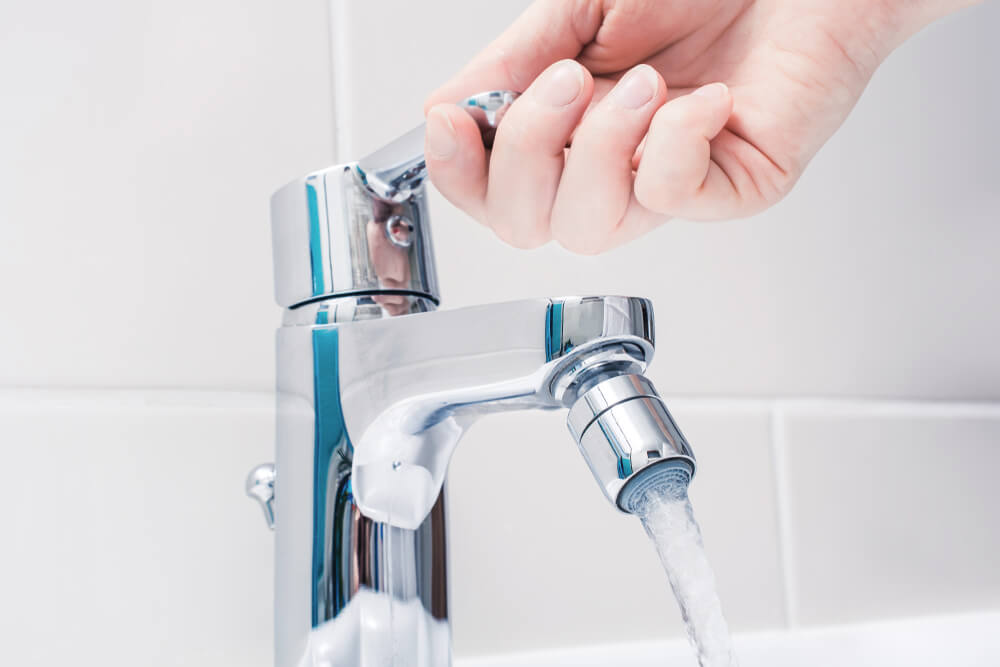 A person's hand is reaching down and opening the metal faucet of a sink in a bathroom. The water is running down from the spout.