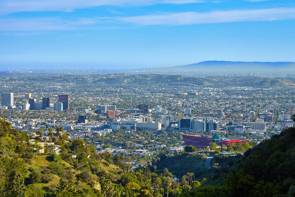 A panoramic view from above of West Hollywood. There are many buildings among the green tress with a deep blue sky above and some mountains out in the distance.