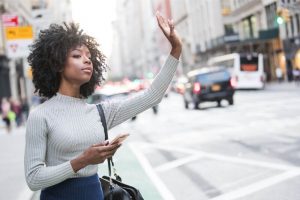 a woman with black hair and a grey shirt is standing on the side of the street while holding her phone and ordering a rideshare