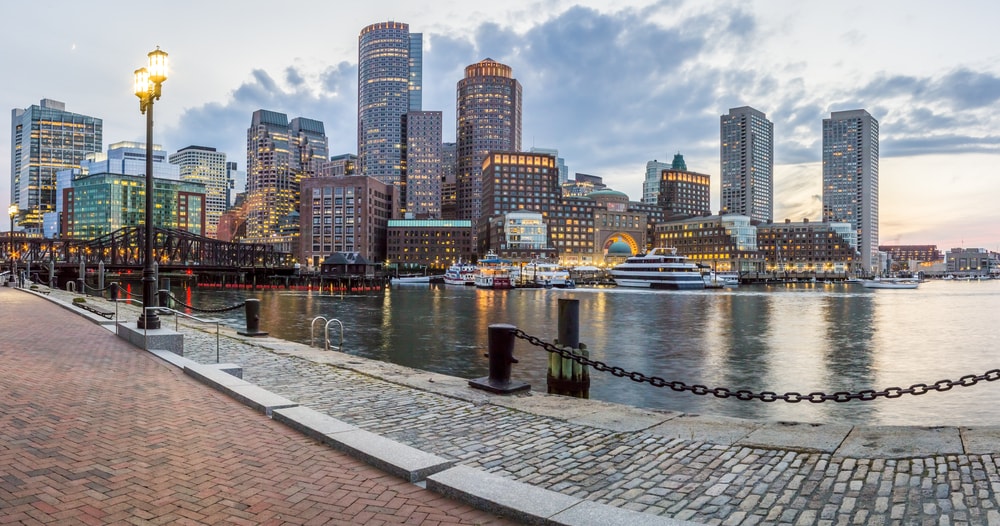 view of the city skyline in the Back Bay neighborhood of Boston. There are many tall buildings across the water with a walkway and a lamp post in the foreground of the photo