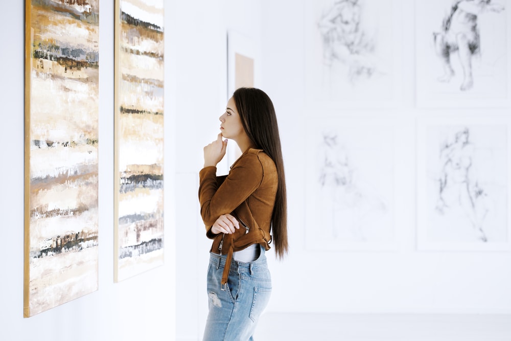 woman wearing a brown shirt and blue jeans stares at a piece of art hanging on a wall in a museum