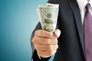 a man wearing a black suit and a maroon tie holds out a fist full of American dollars in cash in front of a blue background