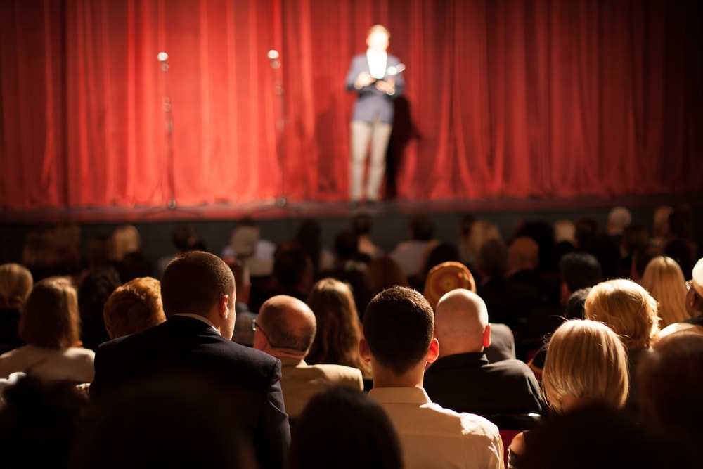 someone standing on a stage in front of a large audience with a red curtain behind them