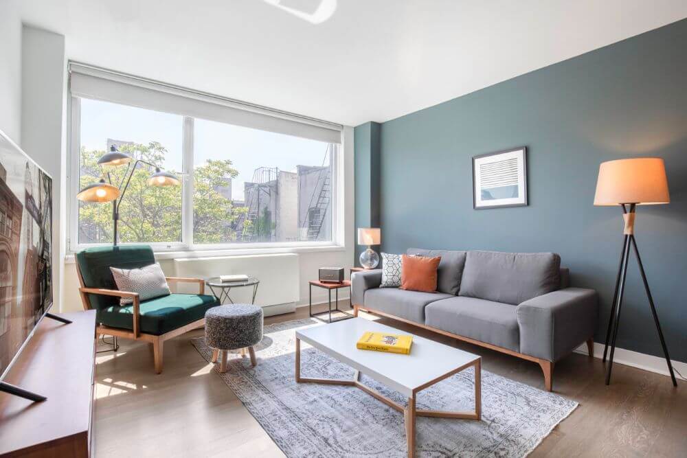 A furnished apartment in New York City that is managed by Blueground. There is a grey couch in front of a dark green accent wall. On the couch are some pillows and in front of the couch is a coffee table with a yellow book. In the corner of the room there is a green velvet chair with a pink pillow.