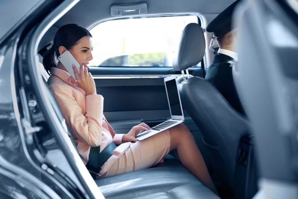 A woman in a light pink outfit sits in the back of a car while holding a laptop on her lap and holding a phone to her ear. There is a driver wearing a hat and a suit jacket sitting in front of her in the drivers seat.