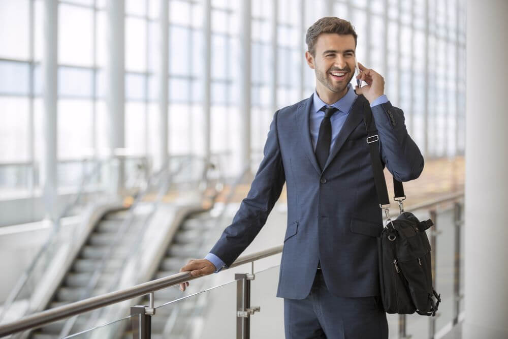 A man in a blue suit and tie with a laptop bag hanging from his shoulder speaks on the phone and smiles. Behind him there are 2 large escalators in the background.