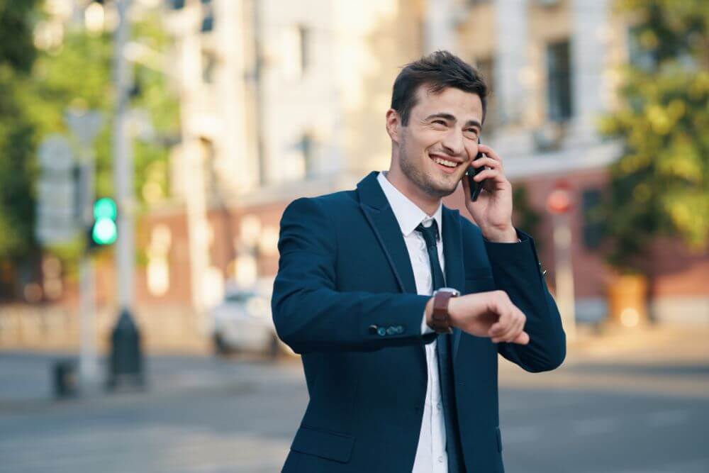 A man wearing a suit jacket and tie looks at his watch and talks on the phone and smiles with a blurry street scene in the background