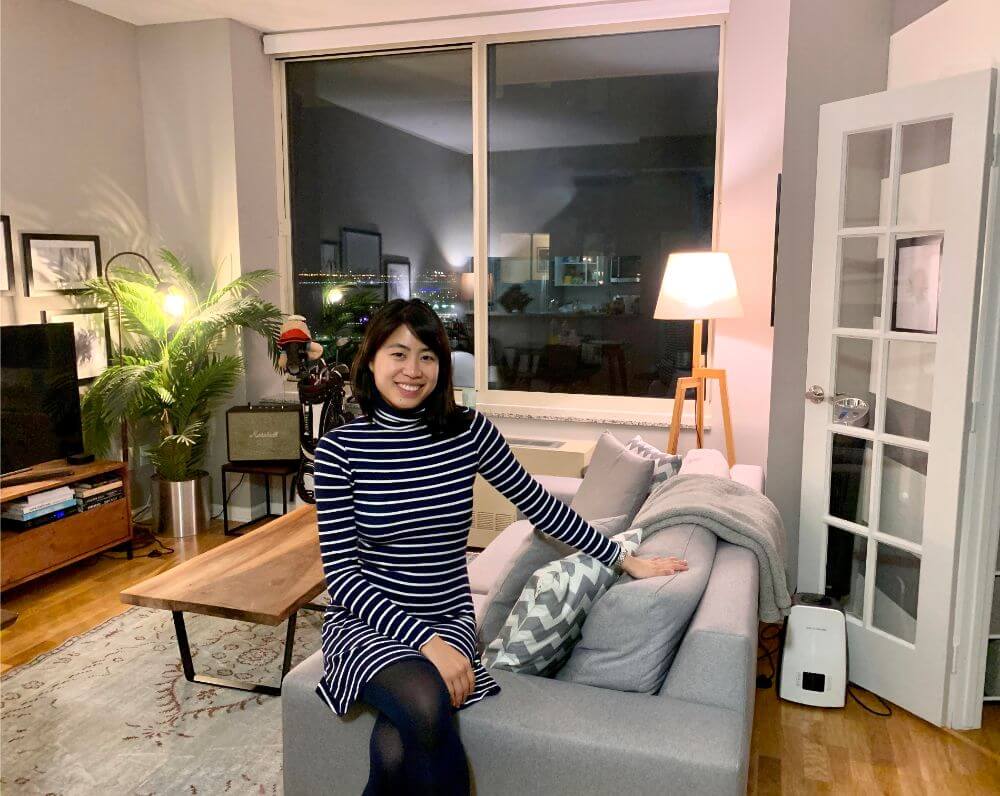 A woman wearing a black and white striped dress is sitting on the armrest of a grey couch in a furnished apartment in Tribeca that is managed by Blueground